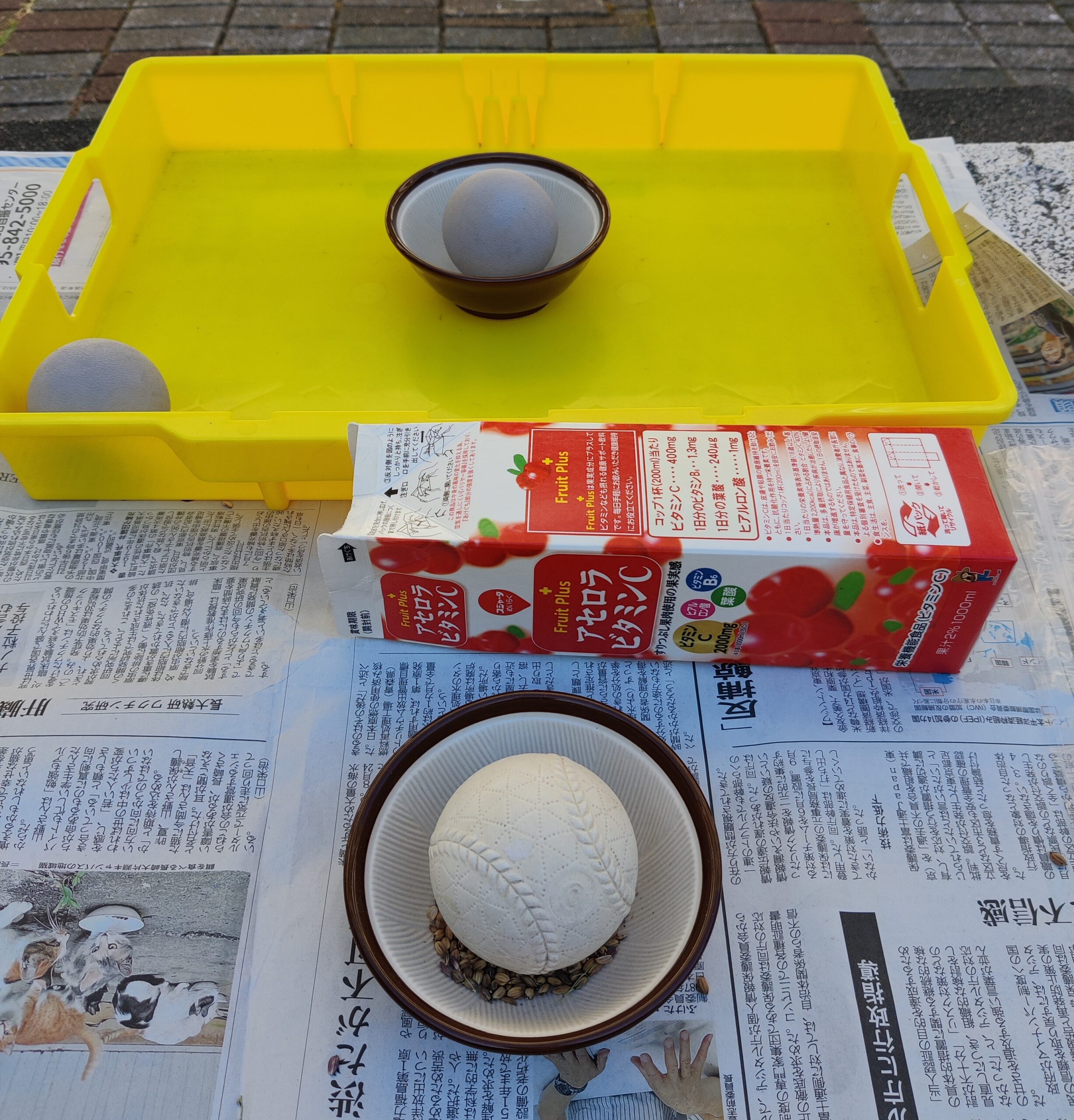 Two mortars with balls in them on a bench. One is in a plastic tray and another one contains rice grains. The bench is covered in newspaper.