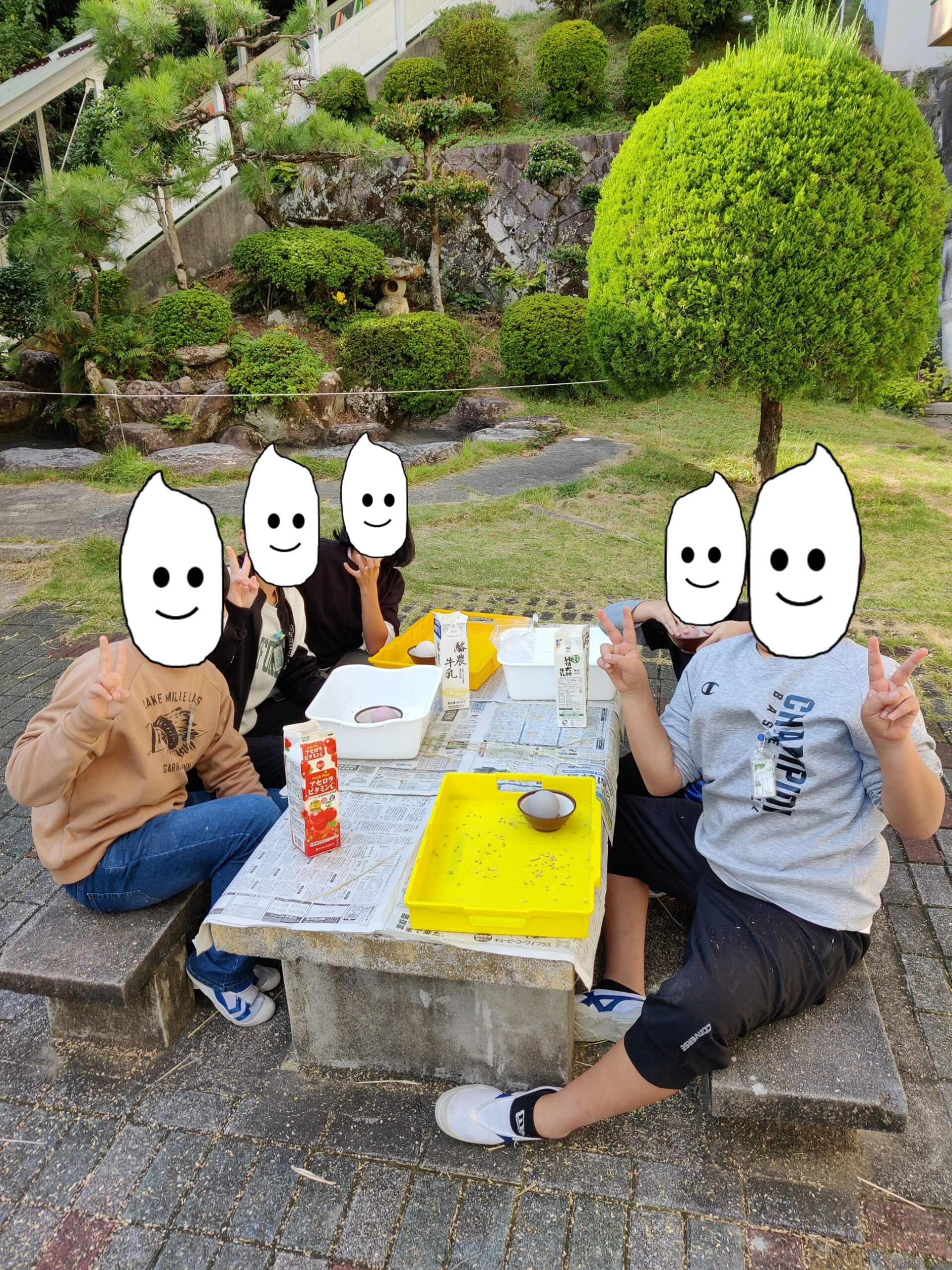 five elementary school children sit at a stone bench and pose for the camera. On the bench there are plastic trays and mortars being used to hull rice.