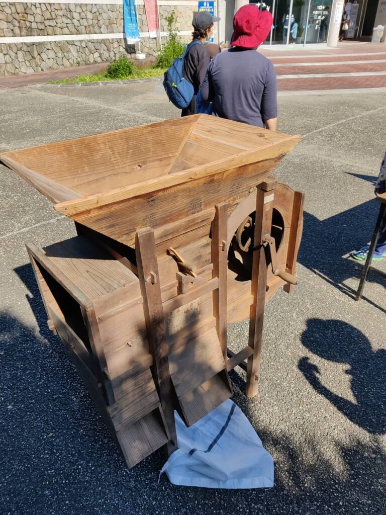 metre tall wooden device has a large open hopper at the top, a handle on the side connected to a cylindrical section. The opposite end to the cylinder there is a a large opening. At the bottom on one side there are two exit chutes.